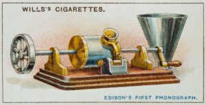 Vintage illustration of Edison's first phonograph, featured on a Wills's Cigarettes trading card. The phonograph consists of a rotating cylinder, a metal horn, and mechanical components mounted on a wooden base, representing one of the earliest sound recording devices.