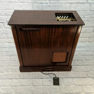 A vintage Wurlitzer Sideman drum machine encased in a wooden cabinet with a dark brown finish. The top panel features various control knobs and buttons, while a wired foot pedal rests on the floor. This early rhythm machine, developed in the 1950s, was one of the first commercially available drum machines.