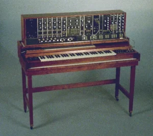 A vintage Moog modular synthesizer mounted on a wooden frame with a built-in keyboard. The synthesizer features numerous knobs, switches, and patch cables on its control panel, reflecting its analog sound design capabilities. This classic instrument, developed by Robert Moog in the 1960s, played a significant role in the history of electronic music.