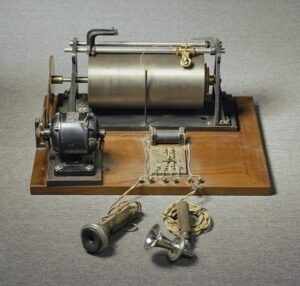 Photograph of the Stahldraht-Telegraphon, an early magnetic sound recording device invented by Valdemar Poulsen in the late 19th century. The machine features a rotating metal wire spool, a wooden base, an electrical motor, and attached microphones, representing a pioneering development in audio recording technology.