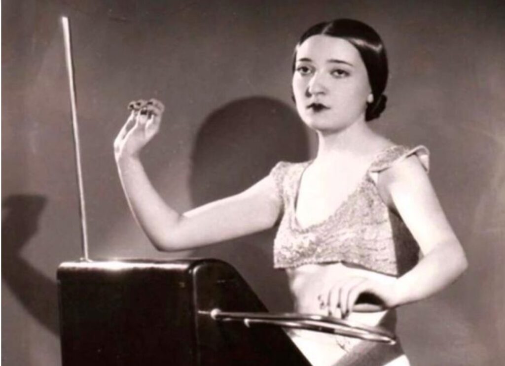 Black and white photograph of Clara Rockmore, a virtuoso theremin player, performing on the electronic instrument. She is elegantly dressed and poised, using precise hand movements to control pitch and volume without physical contact. The theremin, an early electronic instrument, features a vertical antenna for pitch and a horizontal loop for volume control. Clara Rockmore was one of the most famous theremin performers of the 20th century