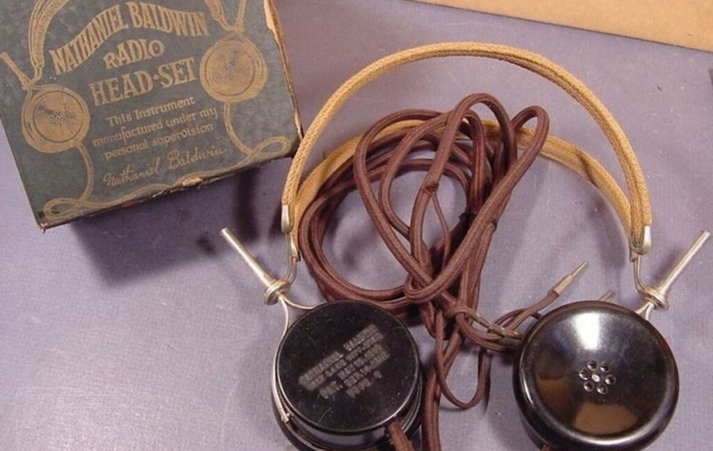 Photograph of an antique Nathaniel Baldwin radio headset from the early 20th century, featuring black earpieces, a fabric-covered headband, and long brown cables. The original packaging is visible in the background, labeled 'Nathaniel Baldwin Radio Head-Set,' highlighting that it was manufactured under Baldwin's personal supervision. This headset was an early innovation in radio communication technology.
