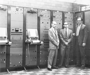 A historic black and white photograph of the RCA Mark II Sound Synthesizer, an early electronic music synthesizer from the mid-20th century. Three men in suits stand in front of the large analog synthesizer system, which features multiple panels with dials, switches, and paper roll input mechanisms.