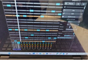 A laptop screen displaying BeepBox 4.2, an online music composition tool. The interface shows a step sequencer with multiple tracks, represented by blue blocks, along with settings such as key (A), tempo (150 BPM), and instrument type (jazz guitar). Various buttons for editing, preferences, and file options are visible on the right. The reflection of a window and surroundings can be seen on the laptop screen.