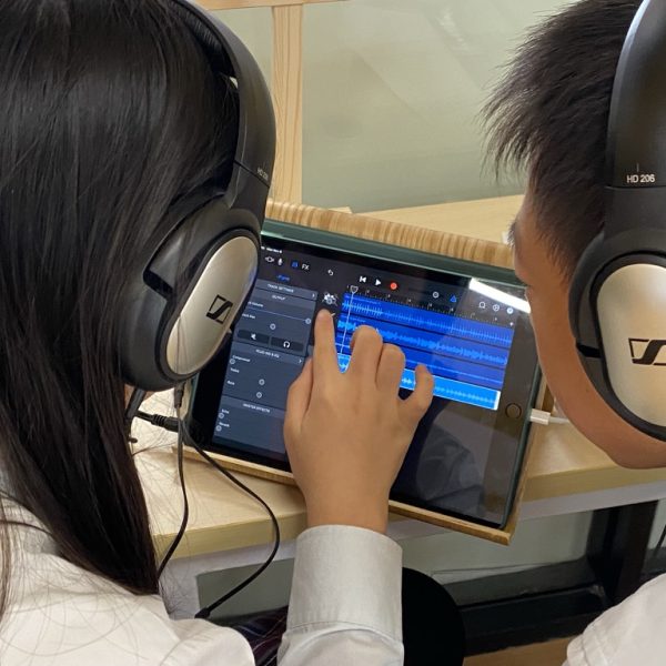 students collaborating on a music project using GarageBand on an iPad. They are both wearing headphones and are focused on the screen as they work with music tracks, likely creating or editing their composition. The interface visible shows the colorful track lanes of GarageBand, indicating they are actively engaged in music creation.