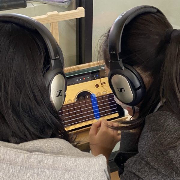 students collaborating on a music project using GarageBand on an iPad. They are both wearing headphones and are focused on the screen as they work with music tracks, likely creating or editing their composition. The interface visible shows the colorful track lanes of GarageBand, indicating they are actively engaged in music creation.